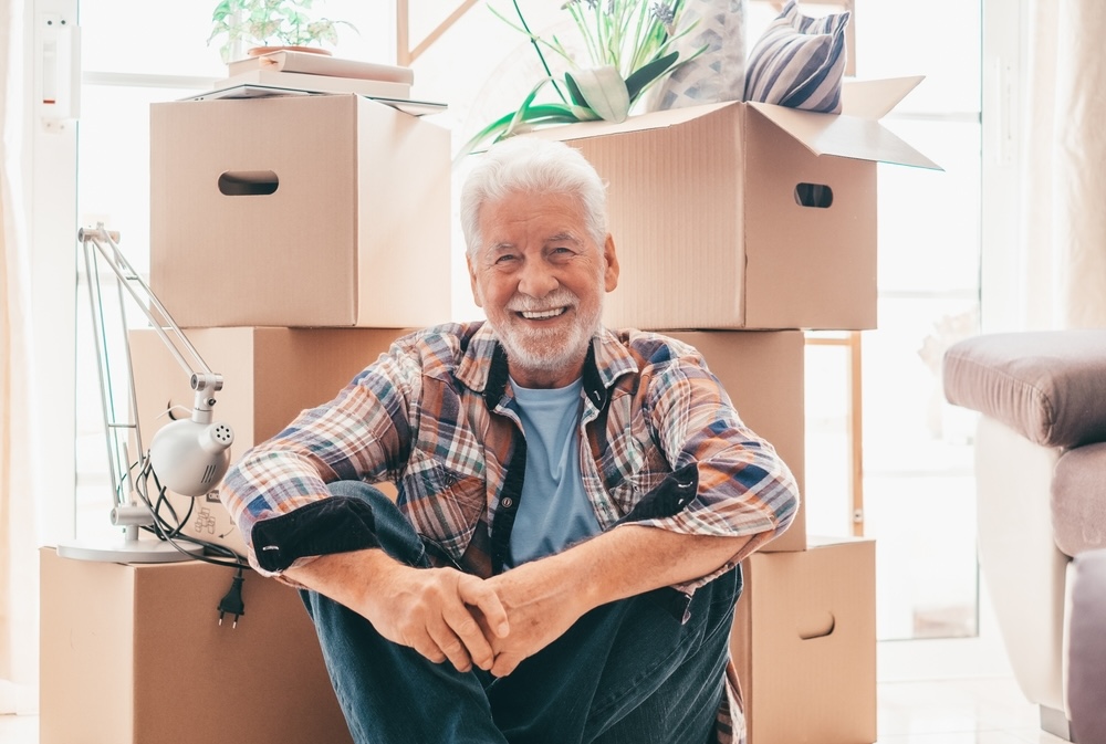 Happy senior man sitting on floor relaxing in new home living room with cardboard boxes packed with office stuff on moving day.